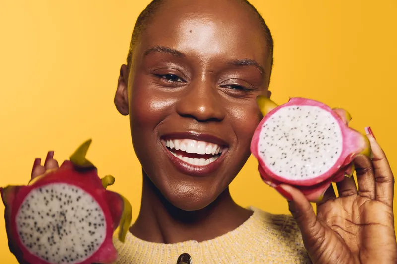 Happy African woman with dark skin holds two halves of dragon fruit on yellow background. Close-up portrait with natural makeup and joyful smile. Concept of healthy lifestyle and fresh fruit.