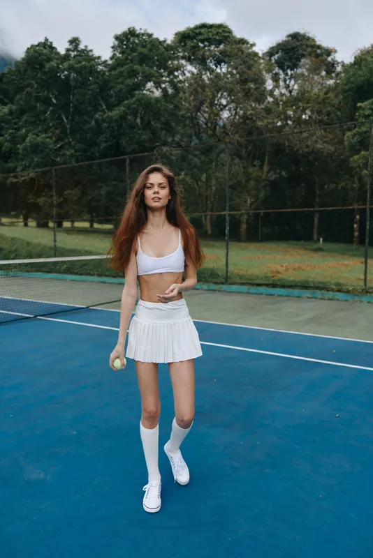 Athletic young woman playing tennis in a white skirt and sports bra, showcasing an energetic expression on a vibrant outdoor court