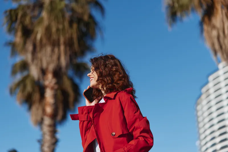A young woman in a red coat stands against a bright blue sky with palm trees behind, smiling as a light breeze moves her hair, exuding confident street style and relaxed mood