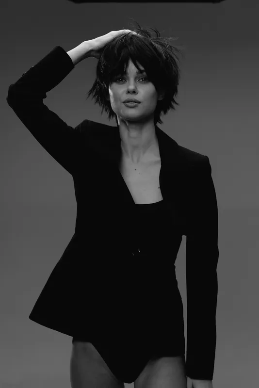 Black and white portrait of a confident woman with short hair wearing a blazer, posing with one hand on her head in a studio setting.