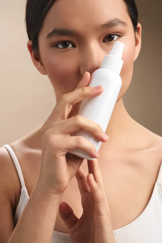 Asian woman applies skincare spray in a clean studio setting, the focus on her face and gentle hands, conveying self care, renewal, and a calm beauty routine.