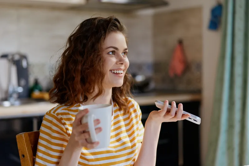 A young woman sits at a cozy kitchen table smiling, holding a mug in one hand and a remote in the other, enjoying a casual morning at home