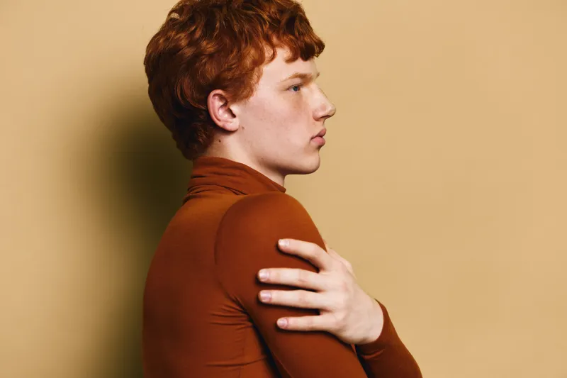 Redhead man in brown turtleneck standing sideways hugging himself with serious expression on beige background. Portrait of thoughtful young adult male with pale skin and curly hair.