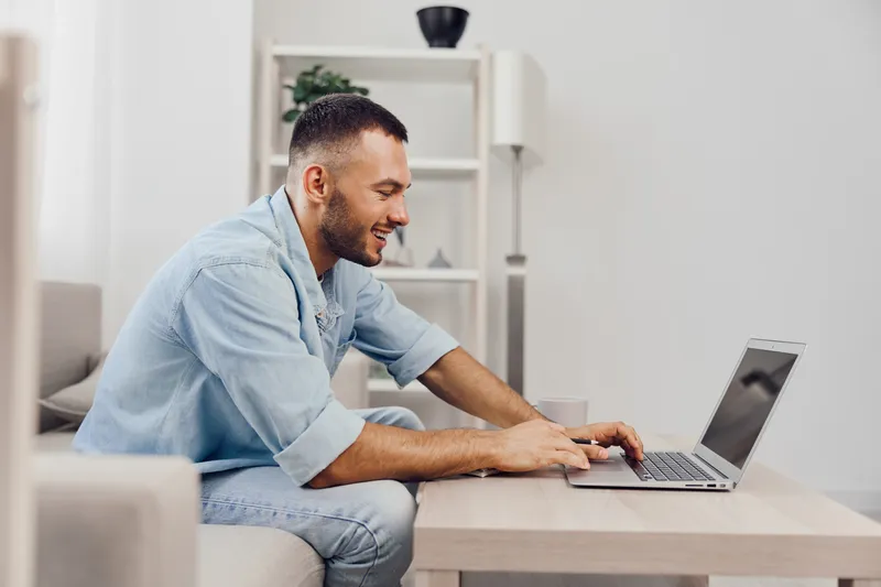 Smiling man working on a laptop at home, enjoying a productive day. Bright space with modern decor, conveying a sense of comfort and focus.