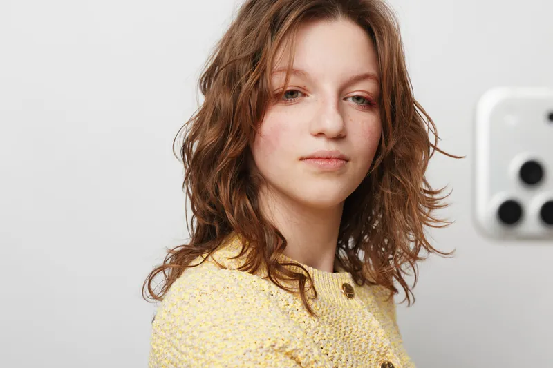 Young woman with curly brown hair wearing yellow sweater looks confidently at camera in bright room. Natural light highlights her clear skin and relaxed expression. Casual portrait concept.