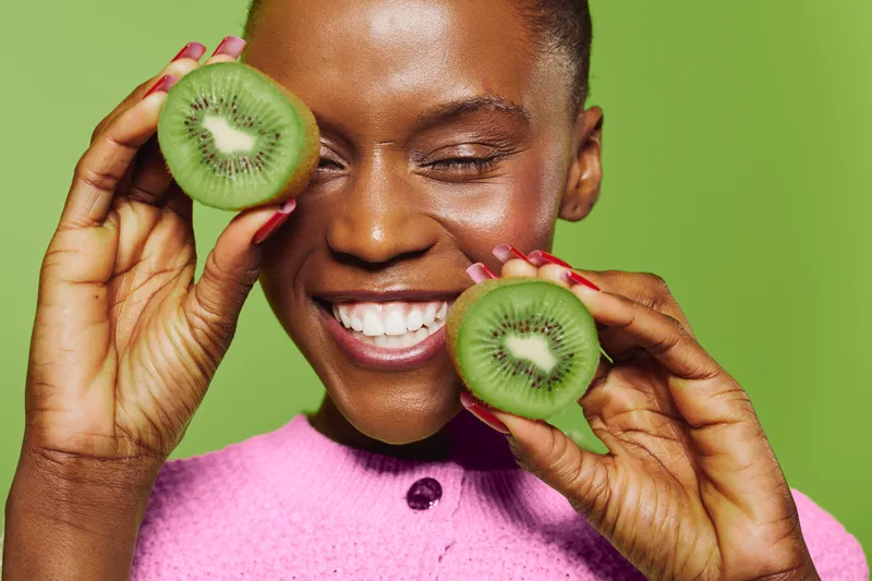 Happy African woman with dark skin holds two kiwi halves near her face smiling with closed eyes. Bright green background and pink sweater create fresh and joyful concept of healthy lifestyle.