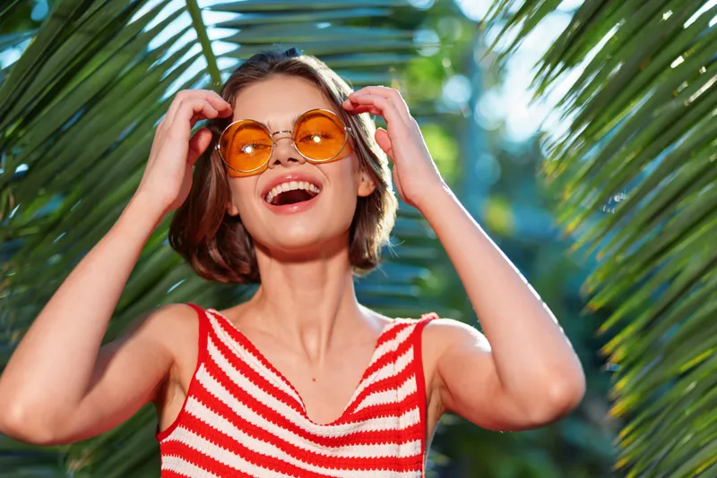 A cheerful woman wearing a red striped top and orange sunglasses smiles brightly while adjusting her frames outdoors, surrounded by tropical greenery and clear blue sky.