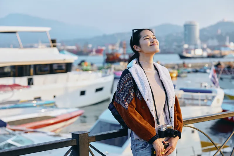 Harbor, boats and a smiling woman in a knit coat and scarf enjoy sunny harborfront scenery by a marina, embracing leisure, fashion and outdoor relaxation under bright skyline light
