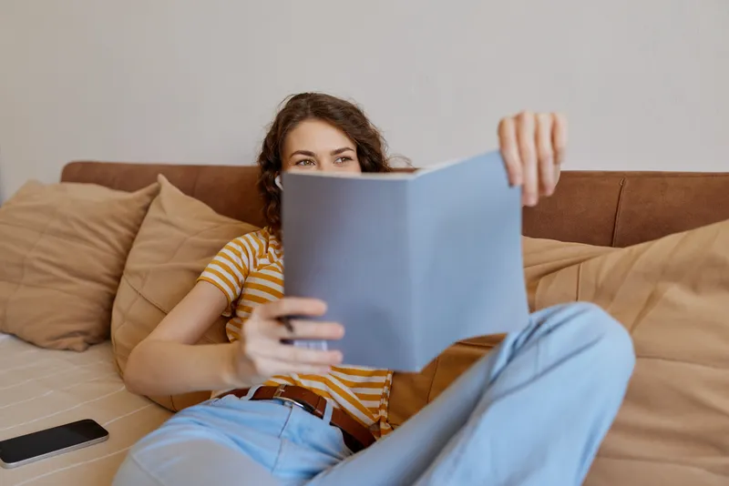 Casual woman lounging on a sofa at home, reading a book in striped shirt and jeans, sunlit room, cozy atmosphere, relaxed lifestyle, intimate moment