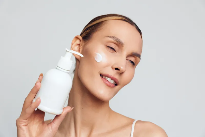 Woman skincare beauty concept, a young woman holds cosmetic bottle and applies cream on face, focusing on hydration and routine, minimal studio background, soft light, calm