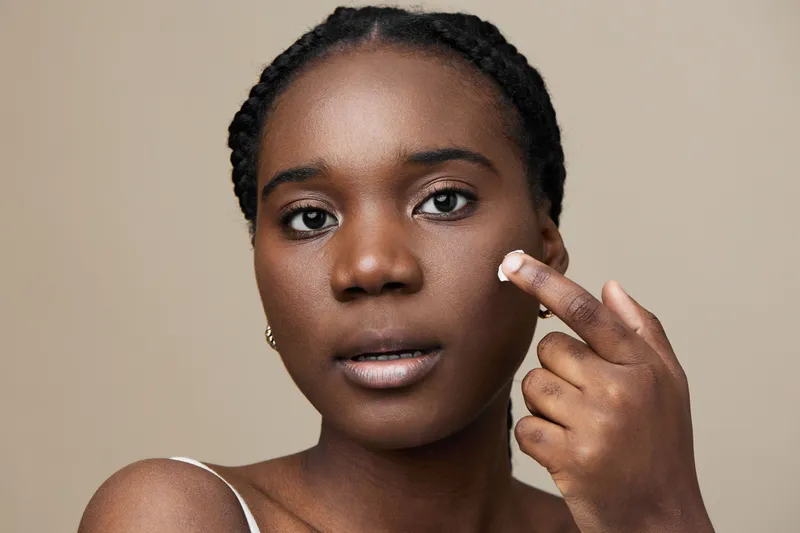 Young Black woman applying skincare product on her face, showcasing healthy skin care routine, neutral background, natural beauty, and confidence in self care