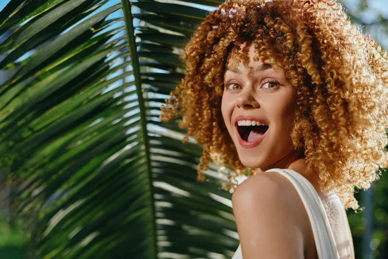 Smiling woman with curly hair enjoys tropical sunlight surrounded by lush green palm leaves, expressing happiness and natural beauty in vibrant outdoor setting.