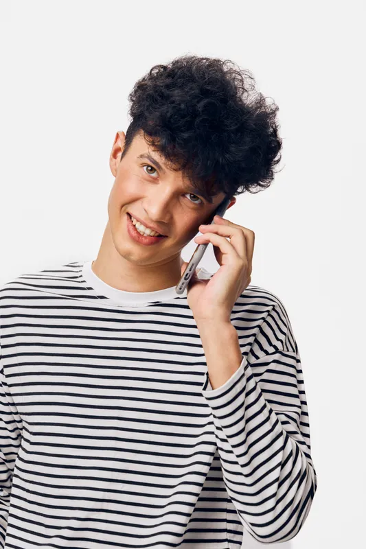 Young man talking on smartphone, wearing black and white striped shirt, smiling and looking at camera, casual style against plain white background.