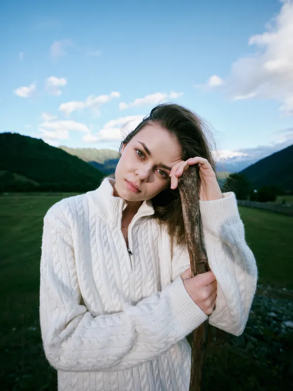 Woman in a white knit sweater stands in a green valley with distant mountains, holding a wooden stick, capturing calm outdoor scenery, rustic charm, and nature lifestyle for travel and wellness themes
