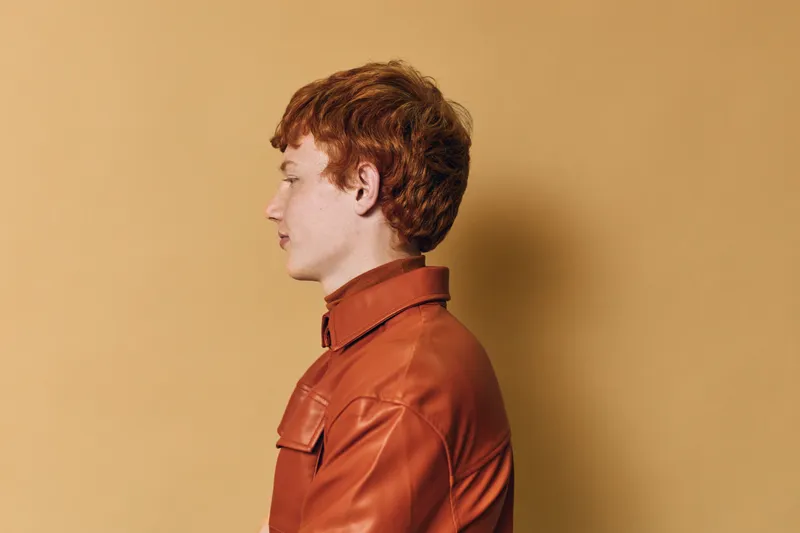 Profile of young man with red hair and freckles wearing orange leather jacket against beige background. Studio portrait with side view of serious male standing still in casual fashion.