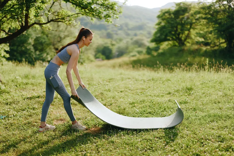 Young woman preparing yoga mat in a green outdoor park for a relaxing yoga session, showcasing fitness, tranquility, and active lifestyle in a serene landscape