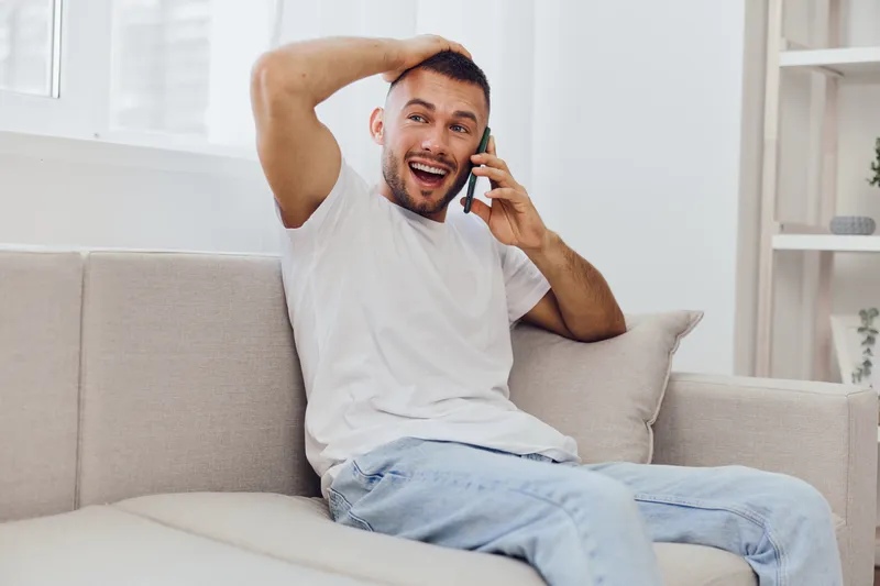 Happy man on the phone, expressing joy and vulnerability, embracing self-care amidst digital distractions and work-life blur in a cozy setting.