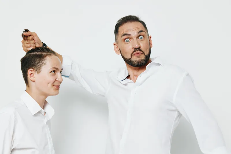 A professional man in a white shirt playfully holds a younger colleague by the hair, both with amused expressions, isolated on a clean white background, conveying humor, lighthearted tension, and