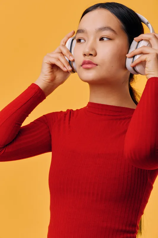 Young woman in red sweater wearing headphones and listening to music with eyes open on orange background. Female portrait, technology and leisure with focused expression in studio.
