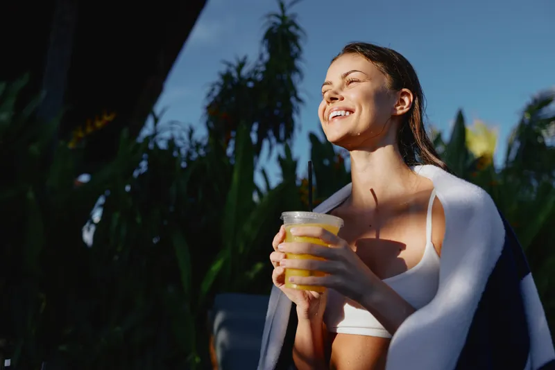 Young woman in a white crop top enjoys a sunny afternoon, smiling while sipping a cold drink on a bright patio, the relaxed vibe amplified by warm natural light.