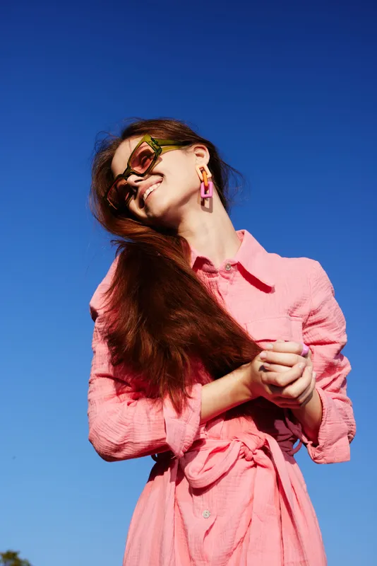 A joyful young woman with long hair, wearing a vibrant pink outfit, enjoying a sunny day against a clear blue sky, exuding happiness and confidence