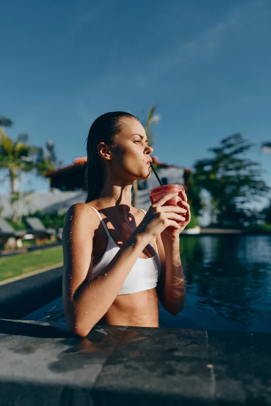 Cheerful woman in a white bikini stands in a sunlit pool, sipping a pink beverage through a straw, enjoying a relaxed outdoor moment by the water and palm shade, with a blue sky overhead