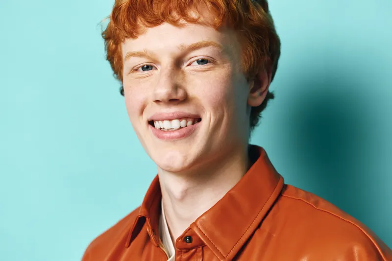 Young man with red hair and freckles smiling in orange shirt on blue background. Portrait, casual style, friendly expression, natural light, confident and happy male model.