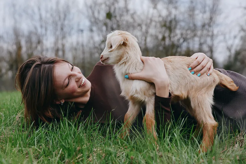Cheerful woman outdoors, cuddling a playful goat kid on lush grass, sunlit moment, relaxed pose, gentle breeze, warm tones, natural colors, countryside backdrop, tender connection