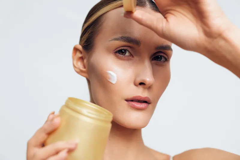 Woman applying face cream with a jar, closeup on white background, hand smooths product on forehead, concept of skincare, selfcare, wellness, soft studio lighting, beauty calm mood