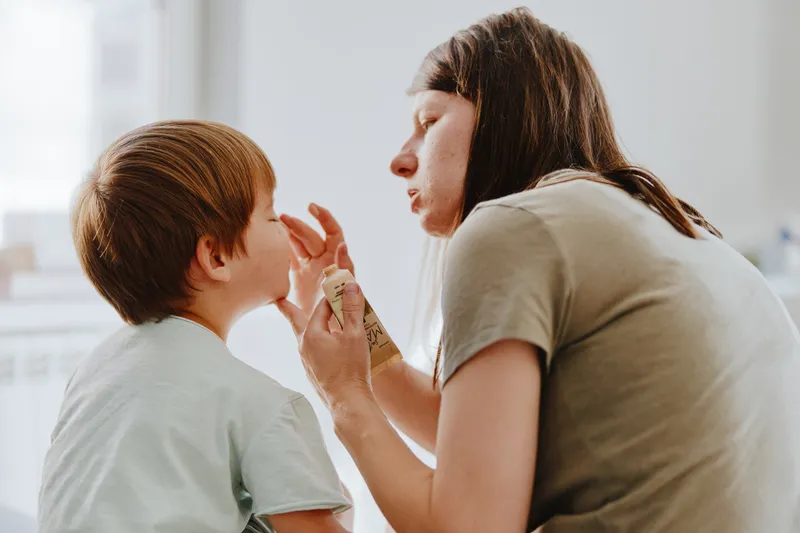 Caring woman applying sunscreen on a child, bright and natural setting, nurturing atmosphere, warm tones, summer vibe, facial expression of focus.