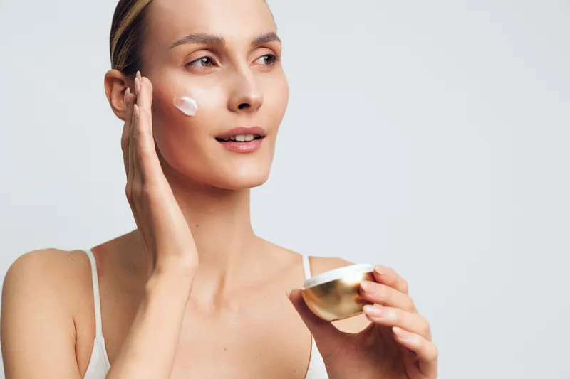 Woman skincare cream beauty face studio applying moisturizer hydration glow, young model gently applies cream to cheek, holding small bowl, calm look, smooth skin, soft light, minimal background
