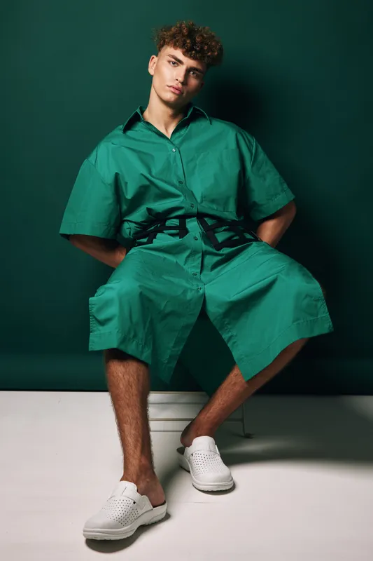 Young man with curly hair wearing green shorts and button-up shirt sitting on chair in studio with neutral air. Casual male fashion model and modern fit clothing outfit portrait with confident pose.