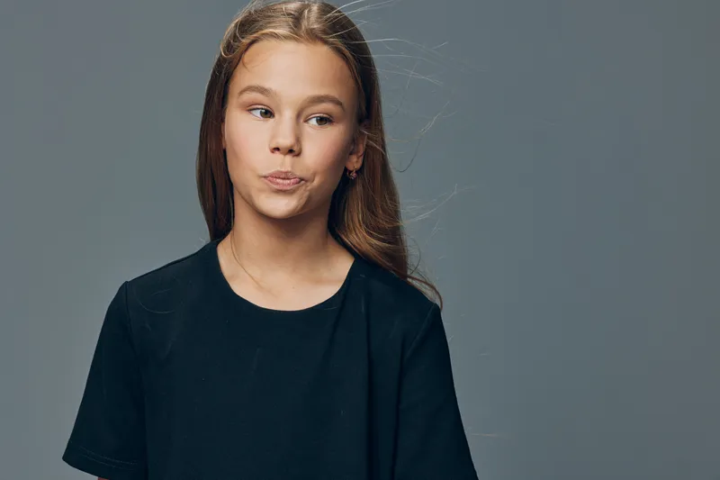 Girl with long hair wearing black t shirt looking confused or thoughtful on gray background. Studio portrait of child with natural expression and casual clothing in neutral space.