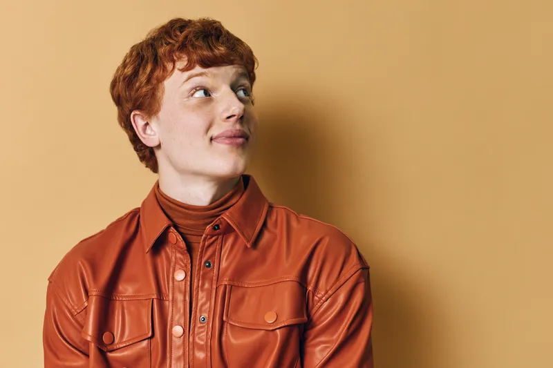 Young man with red hair wearing brown leather jacket and turtleneck looking sideways against beige background in studio portrait.