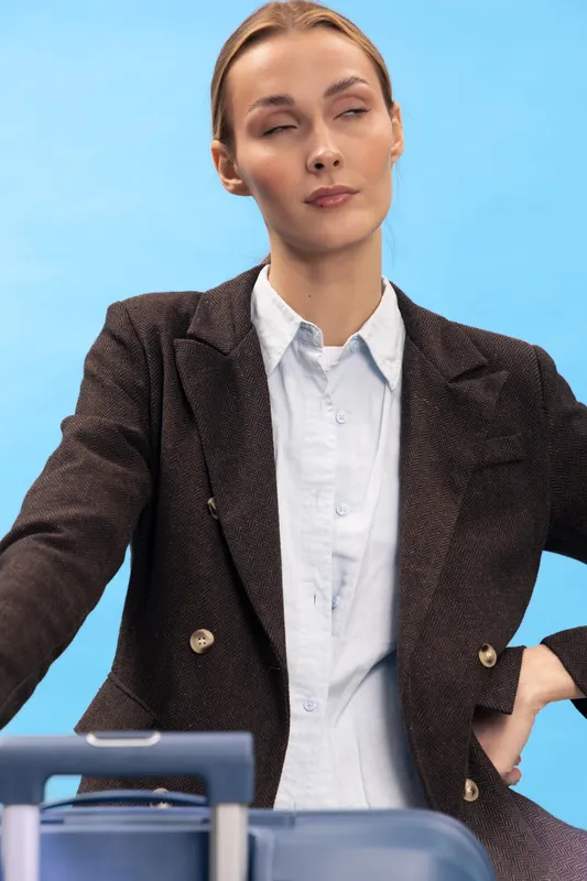 Tired woman in brown blazer and white shirt stands with closed eyes near blue suitcase on blue background. Concept of travel fatigue, business trip stress, and exhaustion before journey.