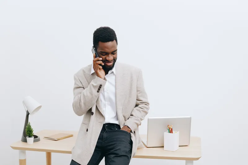 Professional young African American man in casual blazer and shirt talking on mobile phone while standing at modern workspace with laptop, desk lamp, and plants. Business communication, work, technolo