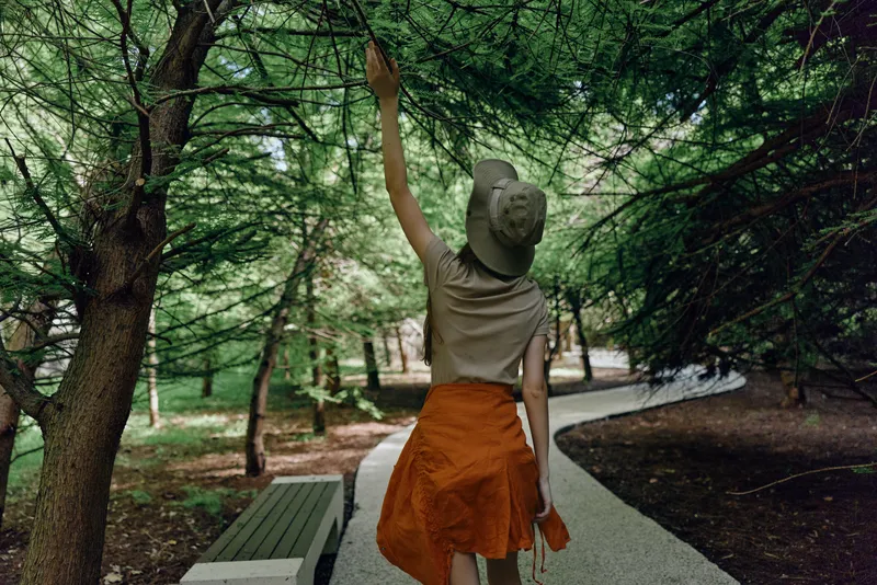 Woman walking in park on a tree-lined path wearing hat and orange skirt, back view reaching up to branches, summer stroll with green trees, bench nearby, casual outdoor leisure in nature.