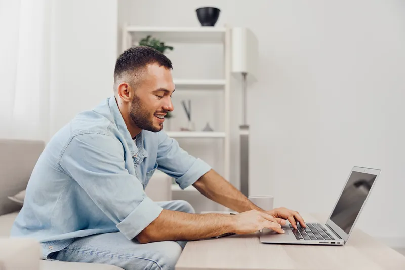 Happy man working on a laptop at home, showcasing work-life blur and soft masculinity. Embracing a digital detox in a cozy environment.