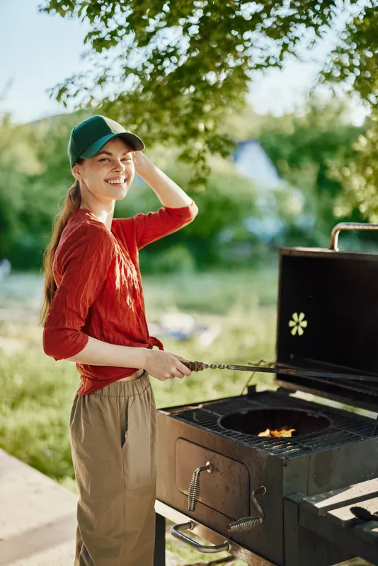 Smiling young woman grilling outdoors in a lush garden, wearing a red shirt and cap, showcasing joy and a love for summer cooking