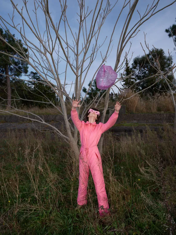 Pink jumpsuit woman outdoors in a field raises arms toward a pink balloon beside a bare tree, playful pose under a blue sky, natural background and whimsical mood