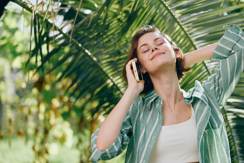 Relaxed young woman outdoors talking on a phone, smiling with eyes closed, surrounded by tropical greenery and warm sunlight.