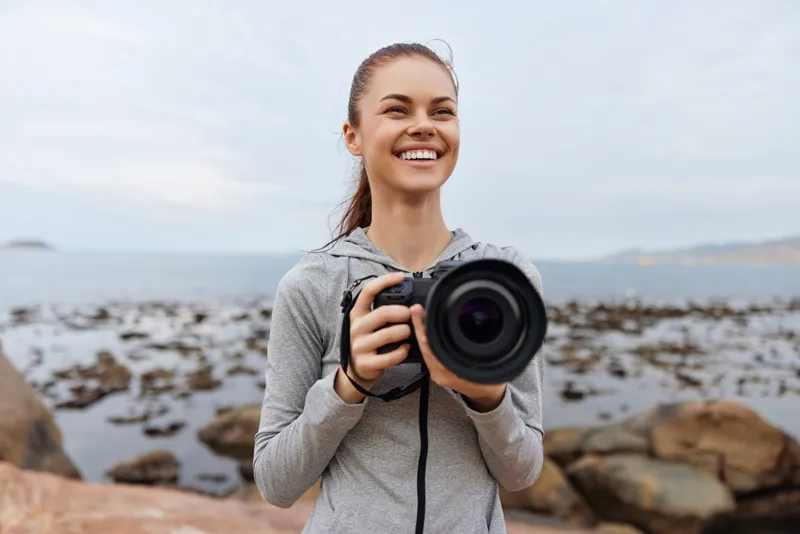 Cheerful young woman in sportswear capturing moments with a camera at a coastal location, showcasing creativity and joy