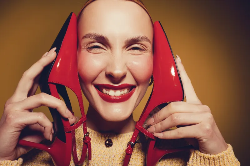 Smiling woman holds red high heels near face with closed eyes and bright lipstick. Warm studio lighting highlights joyful expression and fashion concept on brown background.