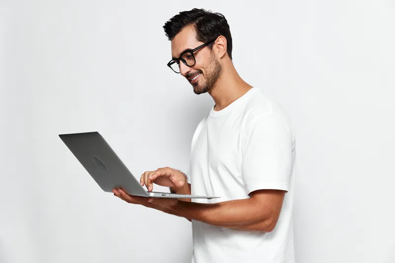 Smiling young man wearing glasses holds a laptop while standing against a plain light background. Engaged in work or leisure, representing productivity and technology.