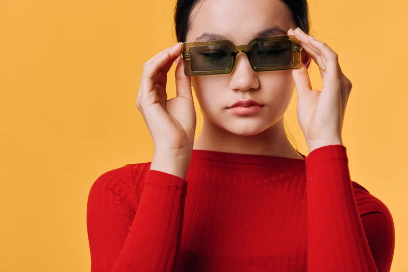 Young woman with sunglasses adjusting eyewear on orange background wearing red long sleeve top. Fashion portrait with casual style and confident expression in studio light.