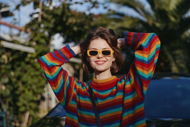 Woman in a colorful striped sweater and yellow sunglasses smiling, young woman posing with hands behind head in tropical outdoor setting, casual outfit for travel and lifestyle fashion.