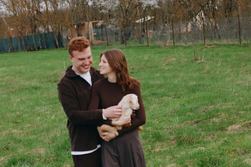 Cheerful couple outdoors, smiling while holding a fluffy puppy, sunlit park moment, relaxed posture, natural colors, lush grass, warm connection, candid interaction, golden hour light