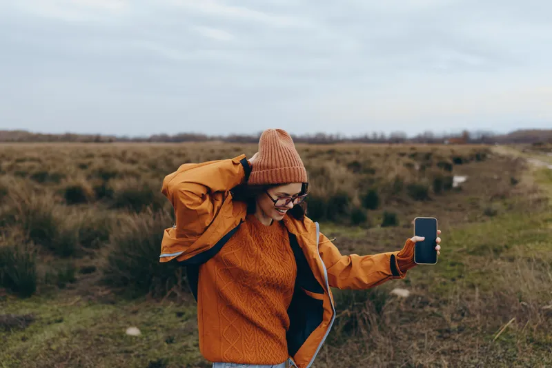 Woman lifestyle outdoors in field landscape, holding smartphone for selfie, wearing orange coat and knit hat, smiling while walking, autumn wind, vast nature background