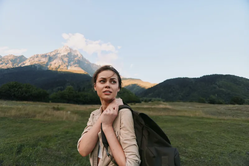 A young woman with a backpack walks through a green meadow as distant mountains rise under a clear sky, symbolizing adventure and exploration.