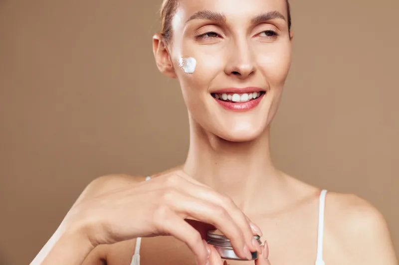 Happy woman applies moisturizer cream on her cheek, showcasing skincare routine, healthy skin and a fresh glow, smiling softly against a neutral background, with soft lighting, serene.
